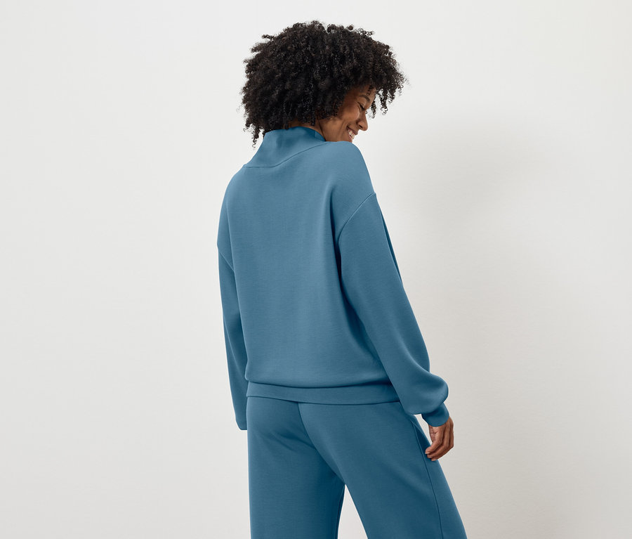 Von hinten ist eine Frau mit dunklen lockigen Haaren in einem blauen Pullover und Hose zu sehen.