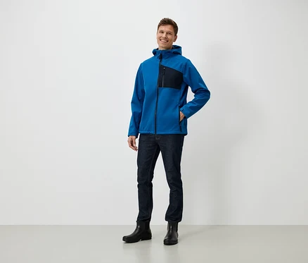 Ein Mann steht und lächelt, er trägt eine blaue Jacke mit Kapuze, Jeans und schwarze Stiefel.
