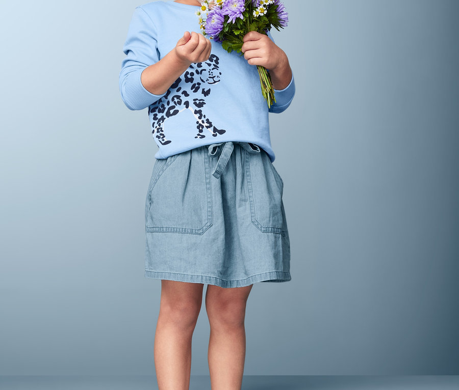 Ein lockiges Mädchen riecht an einem Strauß lila und weißer Blumen. Sie trägt ein blaues Geparden-T-Shirt und einen Baumwoll-Denim-Rock.