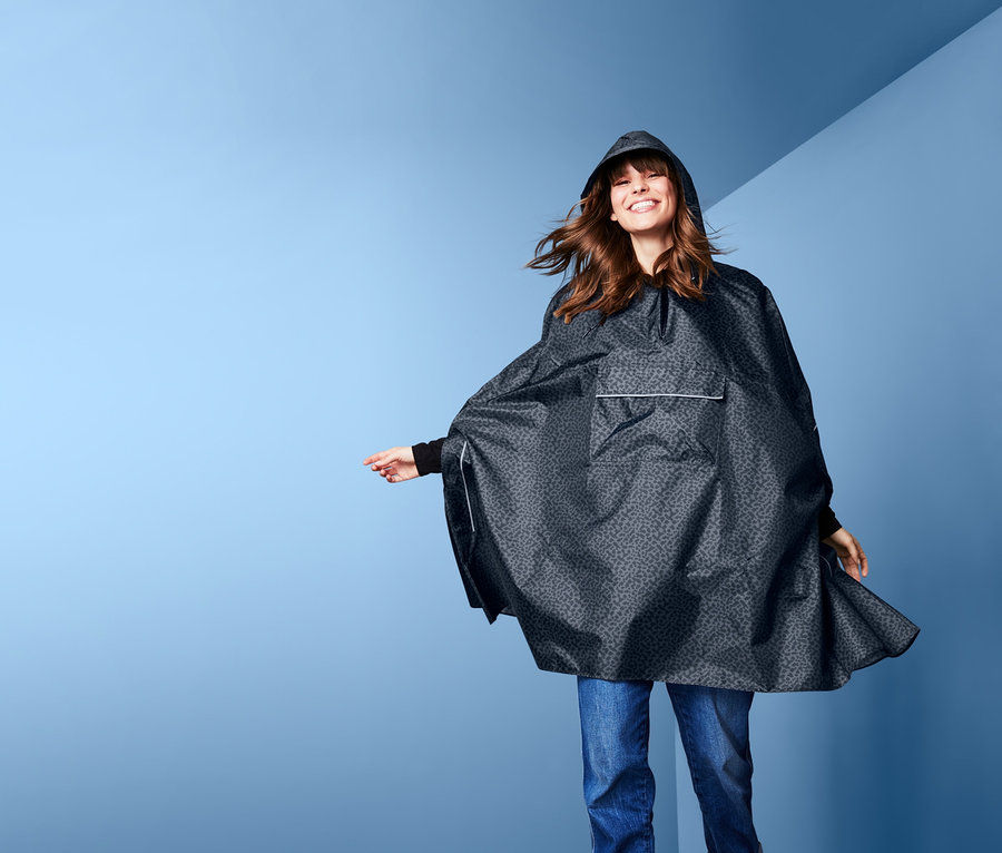 Lächelnde Frau trägt einen grauen Regenponcho mit Kapuze, Jeans und Stiefel.