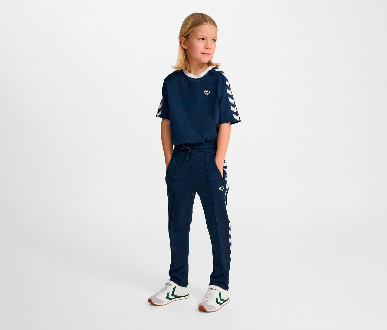 Ein junges Mädchen posiert in einem marineblauen HUMMEL HMLJR Archive Regular Poly Pants Trainingsanzug mit weißen Details.