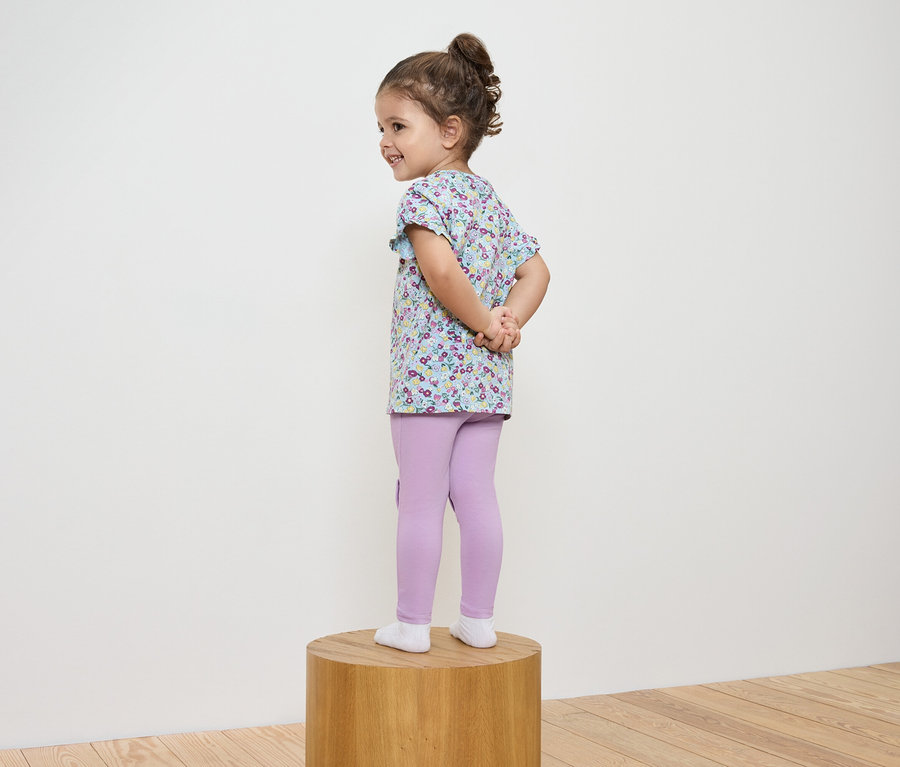 Mädchen in geblümter Bluse und lila Leggings steht auf einem Holzsockel.