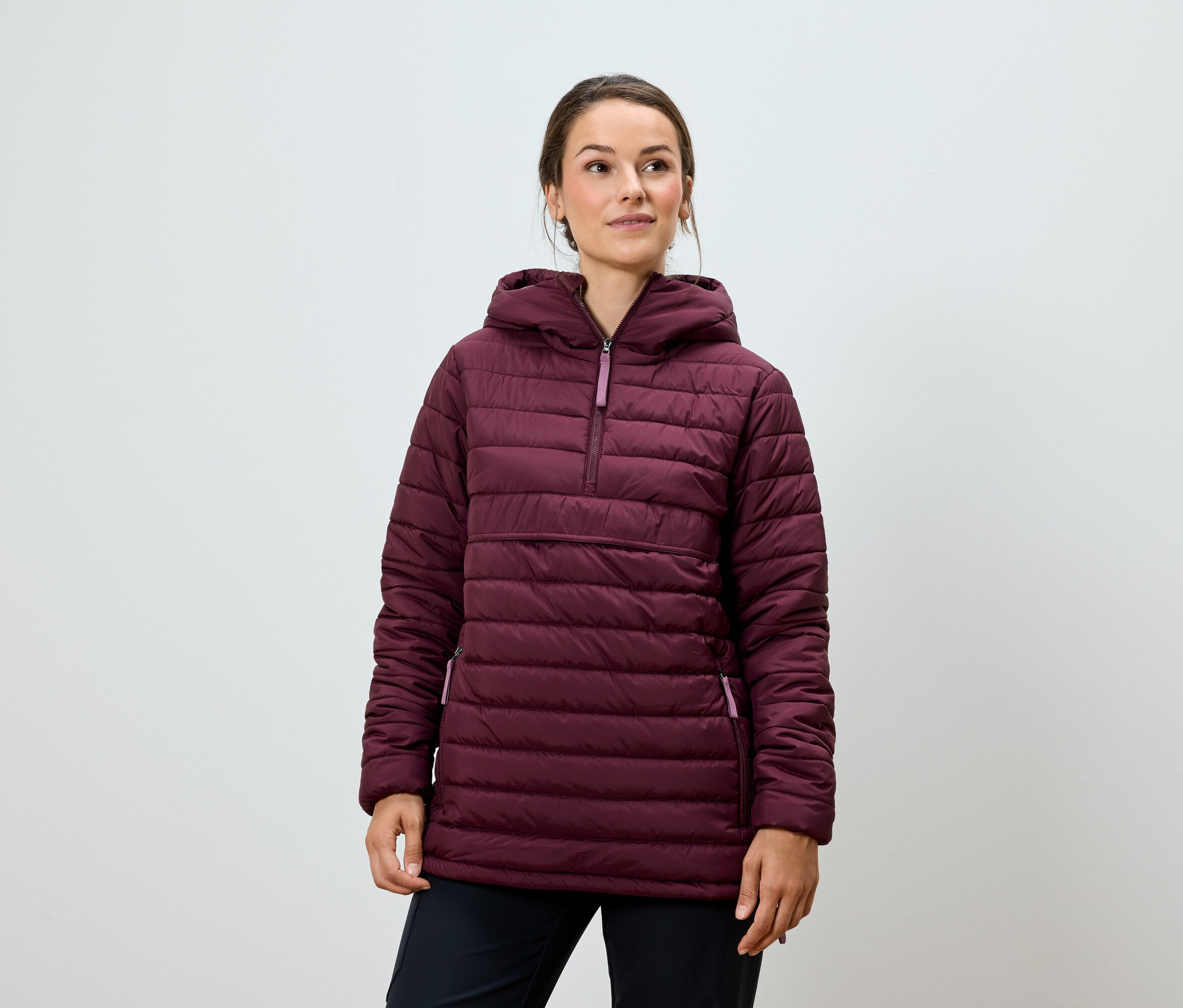 Eine Frau posiert in einer burgunderfarbenen Steppjacke mit Kapuze und einer schwarzen Hose.