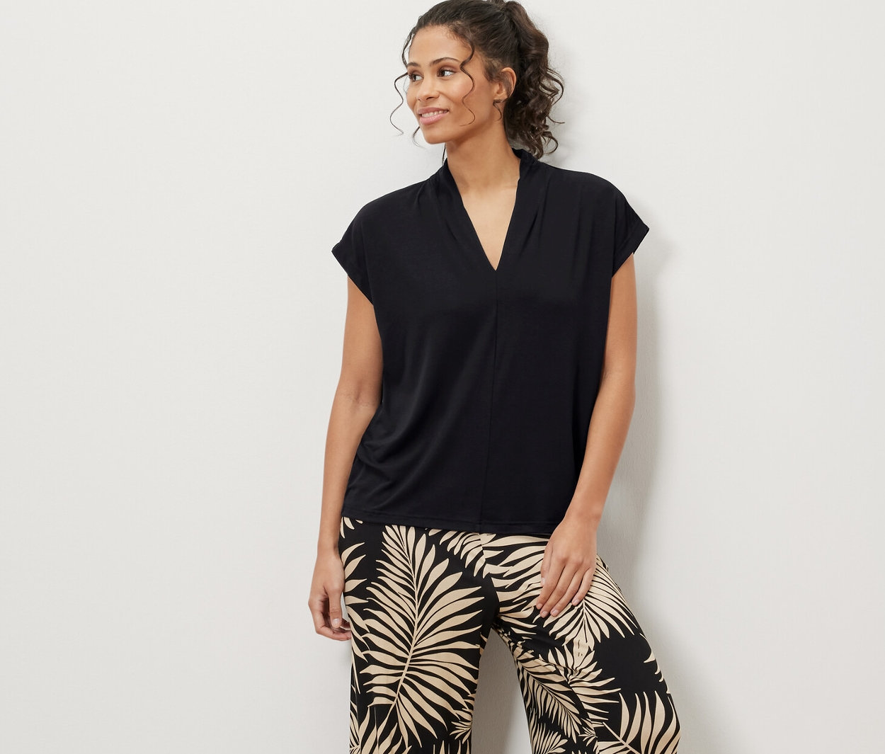 Frau steht und trägt ein schwarzes Blusenshirt mit V-Ausschnitt und Hose mit Blättermuster.