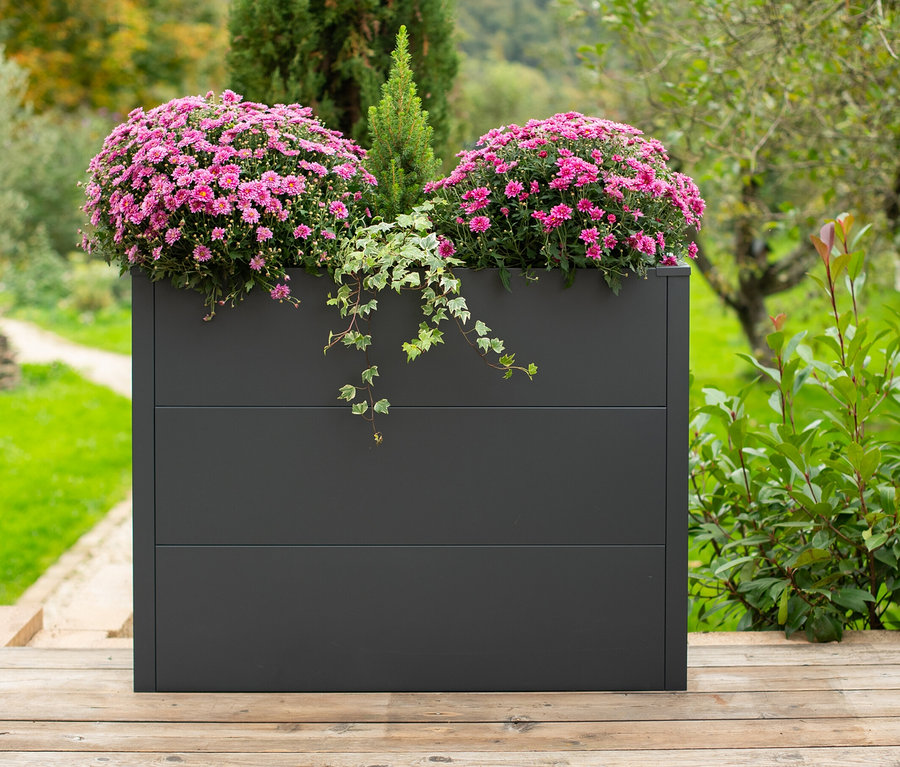 Grauer Pflanzkasten mit rosa Blumen und grünen Pflanzen auf einer Holzterrasse. Im Hintergrund ist ein Garten zu sehen. Produkt: Pflanzkasten mit Einlegeboden, ca. 77 cm