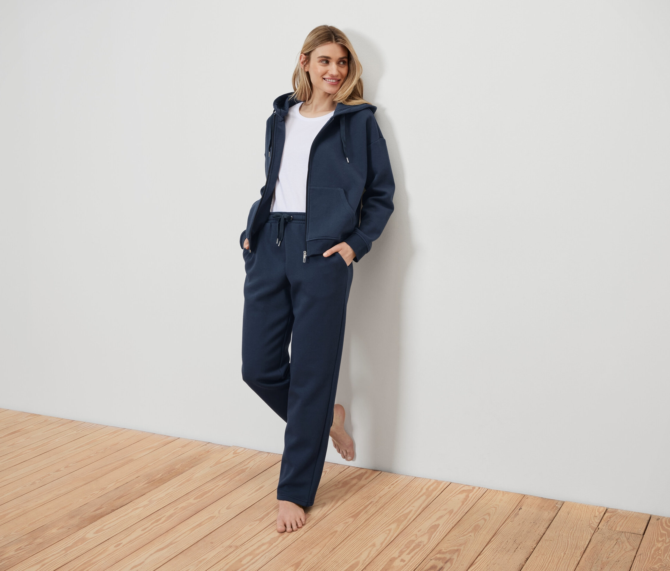 Frau in dunkelblauem Trainingsanzug, an eine weiße Wand gelehnt.