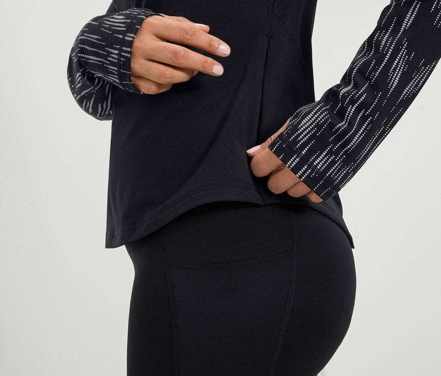 Frau greift in die Seitentasche eines schwarzen, langärmligen Sportoberteils und einer schwarzen Leggings.