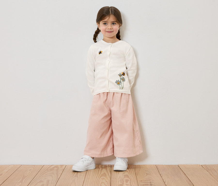 Mädchen mit Zöpfen trägt eine cremefarbene Strickjacke mit Blumen- und Bienenstickerei, eine rosafarbene Hose und weiße Turnschuhe vor einer weißen Wand.