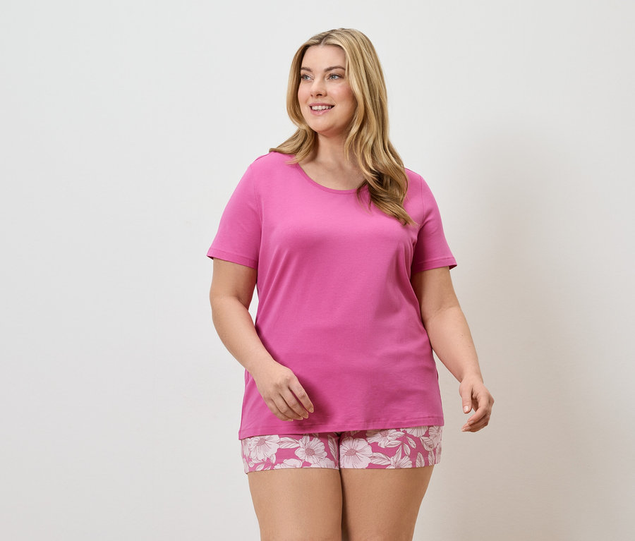 Frau mit blonden Haaren trägt ein pinkes T-Shirt und Shorts mit Blumenmuster vor einem weißen Hintergrund.