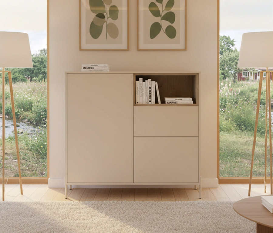 Ein »Hygge« Highboard mit 2 Türen in Kaschmir-Weiß steht zwischen zwei Fenstern mit Landschaftsblick. Darüber hängen zwei gerahmte Bilder.