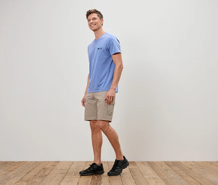 Ein Mann geht auf einem Holzboden und trägt ein blaues T-Shirt, kurze, beigefarbene Hosen und schwarze Turnschuhe.