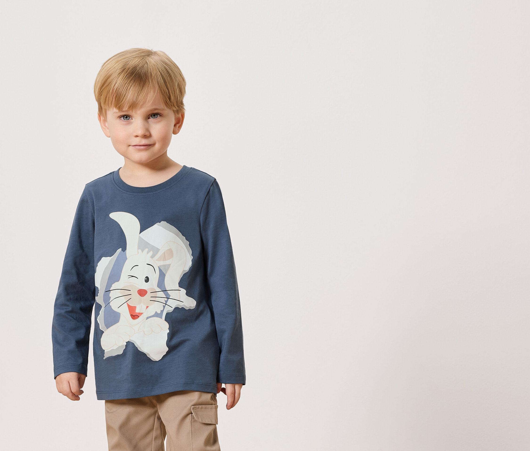 Ein kleiner Junge trägt ein blaues Langarmshirt mit einem weißen Hasenaufdruck und eine beigefarbene Hose.