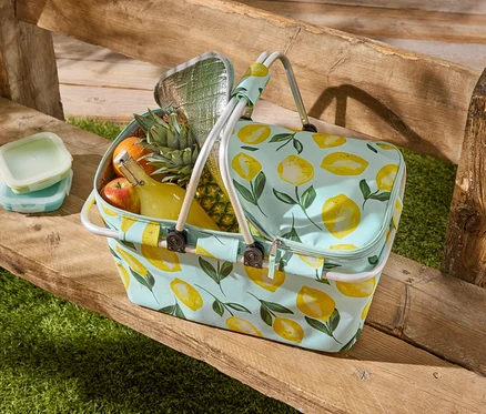 Eine Picknicktasche mit Zitronenmuster steht auf einer Holzbank, gefüllt mit Obst, einer Flasche und zwei Frischhaltedosen.