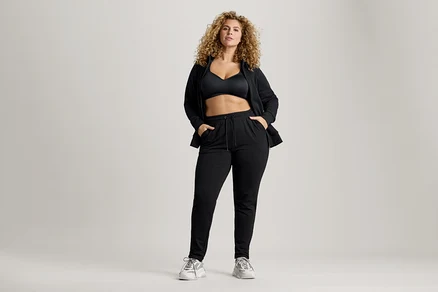 Frau mit lockigem Haar, die einen schwarzen Sport-BH, eine Jacke und eine Jogginghose trägt und die Hände in den Taschen hat.