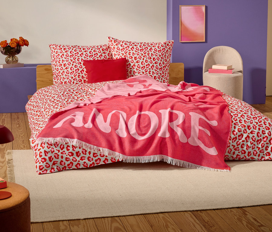 Auf einem Bett mit rot-weißem Leopardenmuster liegt eine rote Decke mit dem Wort "AMORE" in großen weißen Buchstaben.