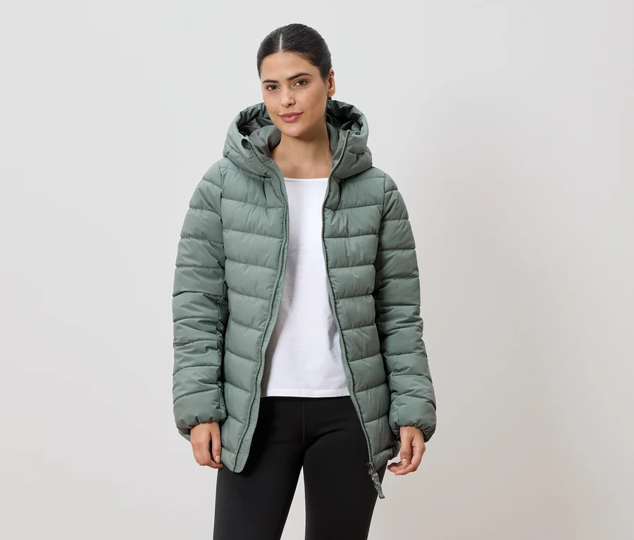 Frau mit Pferdeschwanz trägt eine hellgrüne, gesteppte Jacke mit Kapuze, ein weißes T-Shirt und schwarze Leggings vor einem weißen Hintergrund.