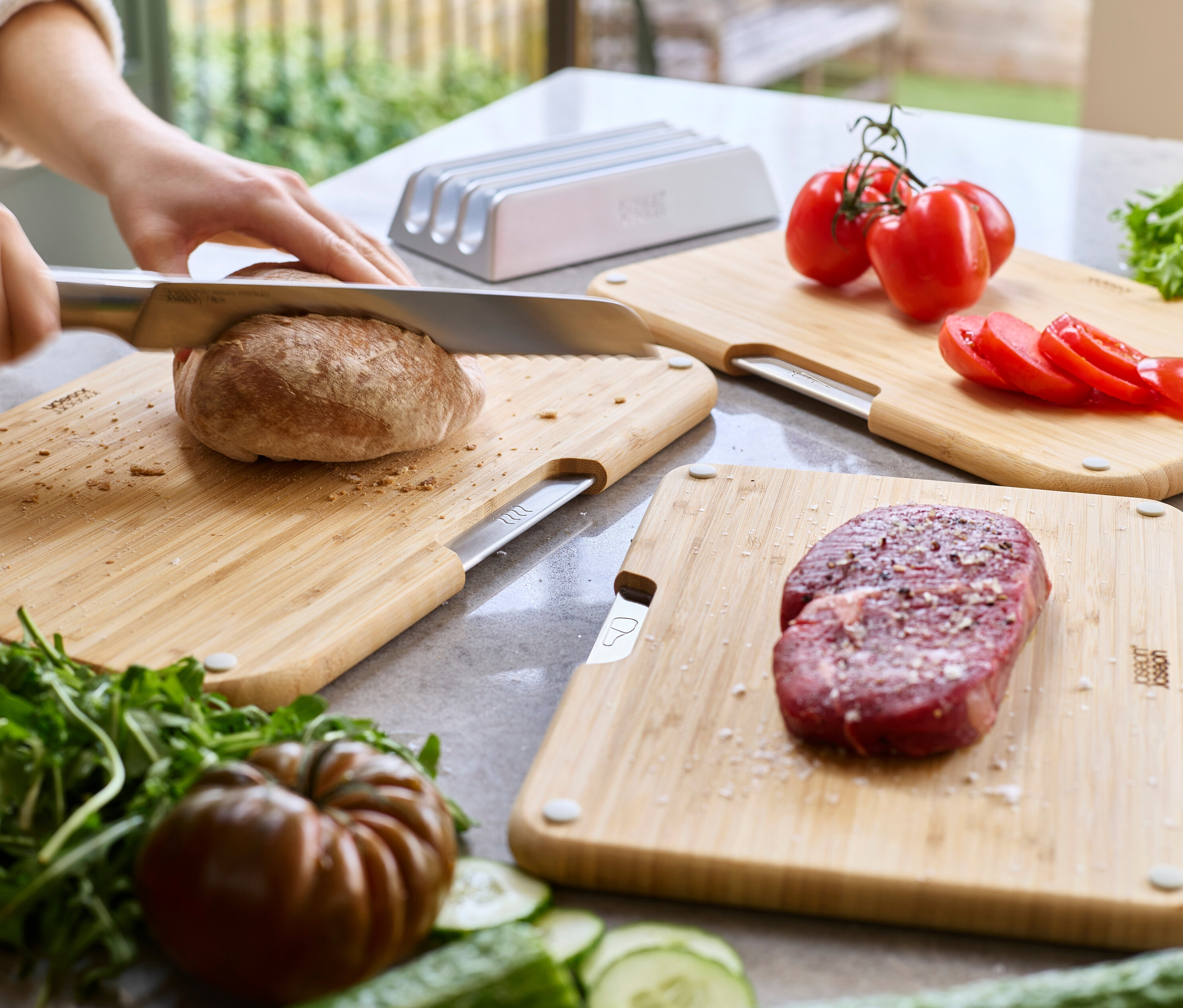 Eine Person schneidet ein Brot auf einem Schneidebrett. Mehrere Schneidebretter mit Tomaten und Fleisch sind ebenfalls sichtbar.