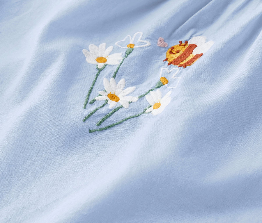 Nahaufnahme eines hellblauen Stoffes mit gestickten Gänseblümchen und einer Biene.