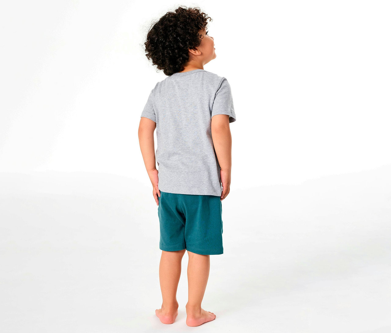 Junge von hinten in kurzem SCHIESSER Kinder-Pyjama aus grauem T-Shirt und grünen Shorts, der nach oben schaut.
