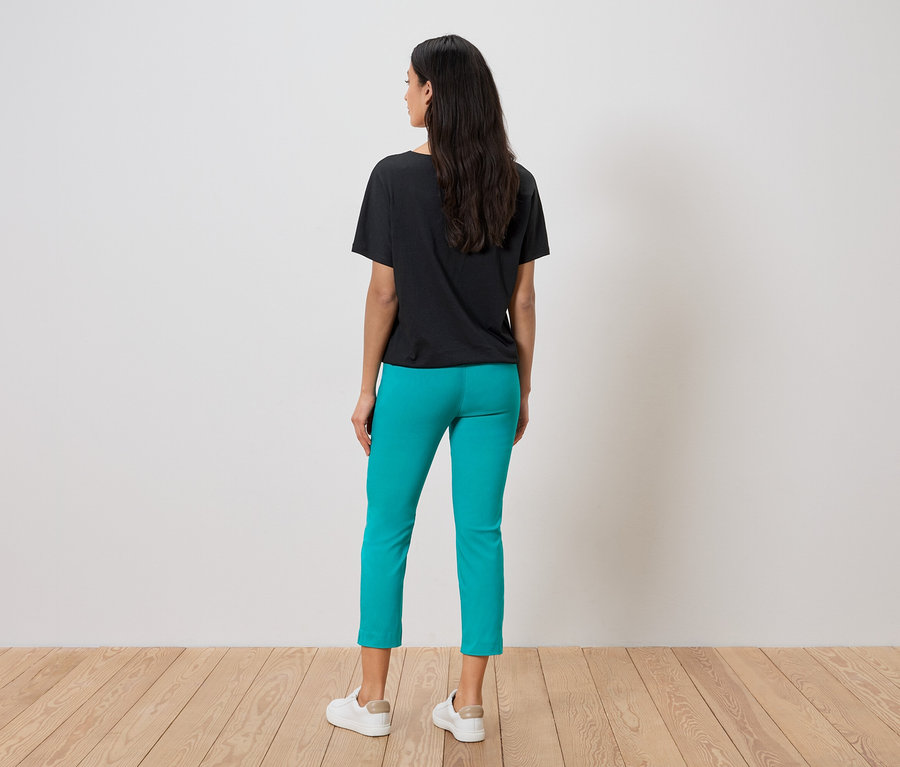 Rückansicht einer Frau mit dunklen, schulterlangen Haaren, die ein schwarzes T-Shirt, eine türkisfarbene Hose und weiße Turnschuhe trägt.