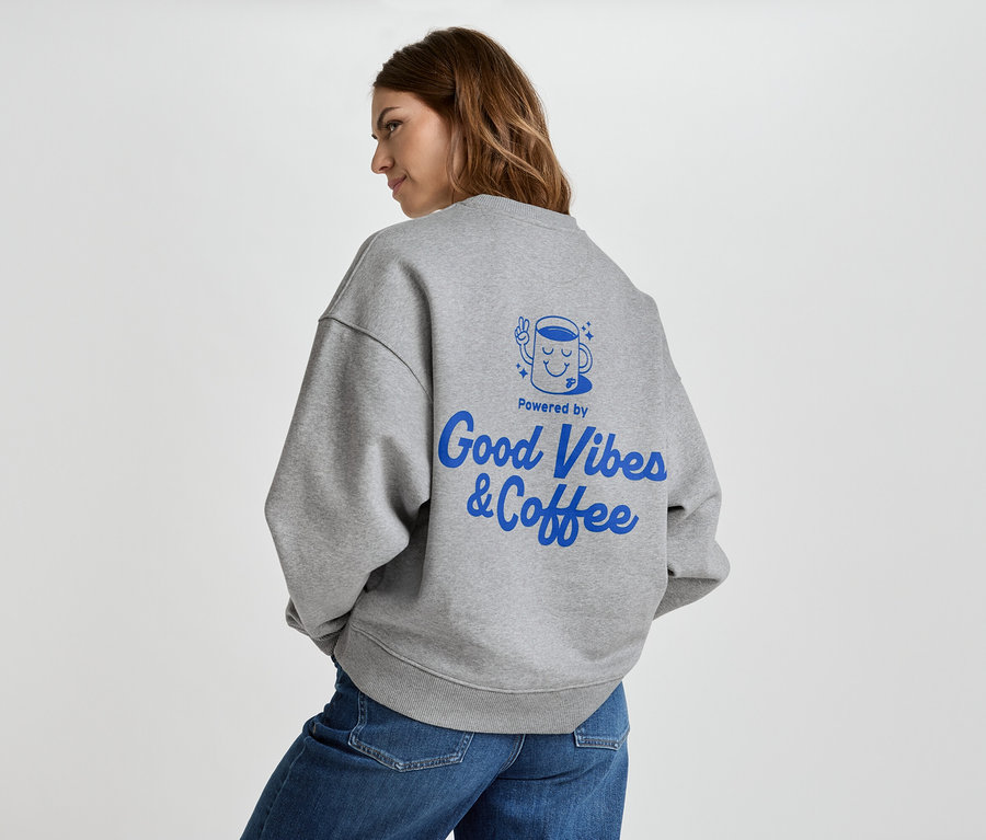 Frau trägt graues Sweatshirt mit blauem Aufdruck "Good Vibes & Coffee" auf dem Rücken, dazu Jeans.