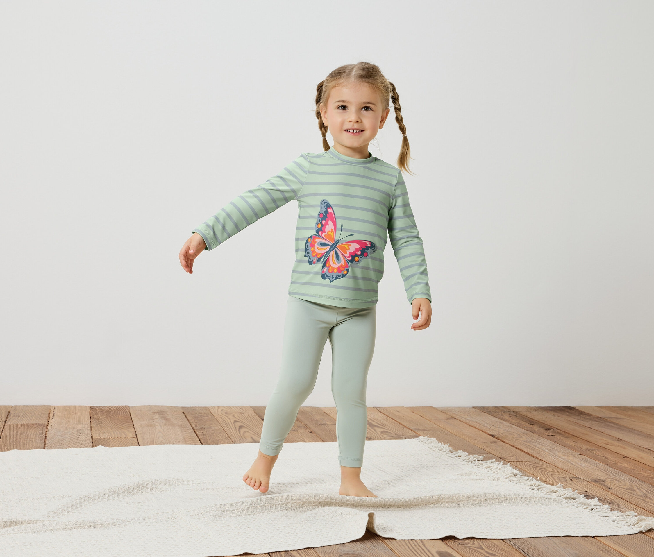 Mädchen mit Zöpfen, trägt ein hellgrünes, gestreiftes Oberteil mit Schmetterlingsaufdruck und Leggings.
