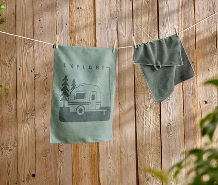 Ein grünes Geschirrtuch mit Wohnwagen-Motiv hängt neben einem strukturierten Handtuch an einer Wäscheleine vor einer Holzwand.