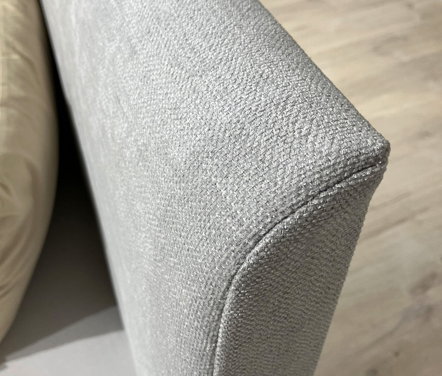 Detailaufnahme eines hellgrauen, stoffbezogenen Möbelstücks, möglicherweise ein Sofa oder ein Sessel, mit einem beigen Kissen daneben.