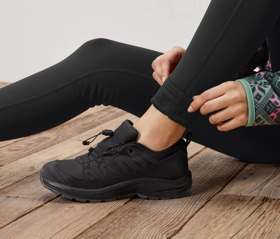 Eine Person sitzt auf einem Holzboden und krempelt ihre schwarzen Leggings hoch, neben einem schwarzen Schnürschuh.