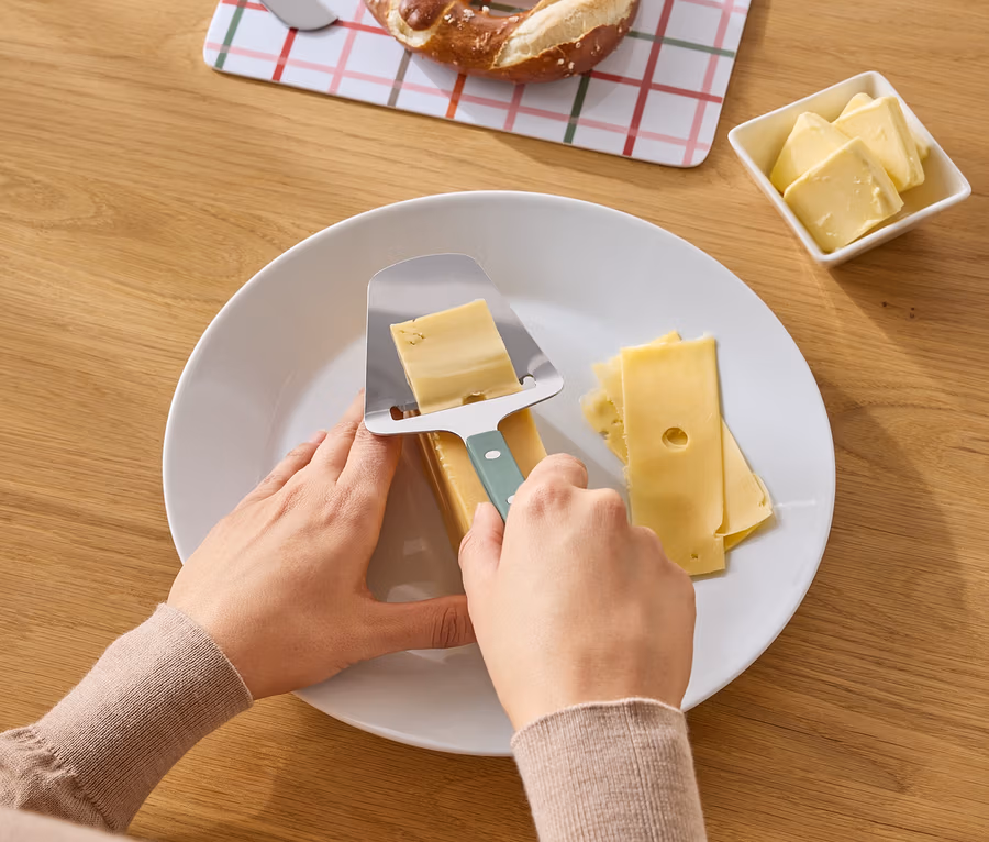 Eine Person schneidet Käse mit einem Käsehobel auf einem Teller. Brezel, Butter und ein Messer liegen auf einem Holztisch.