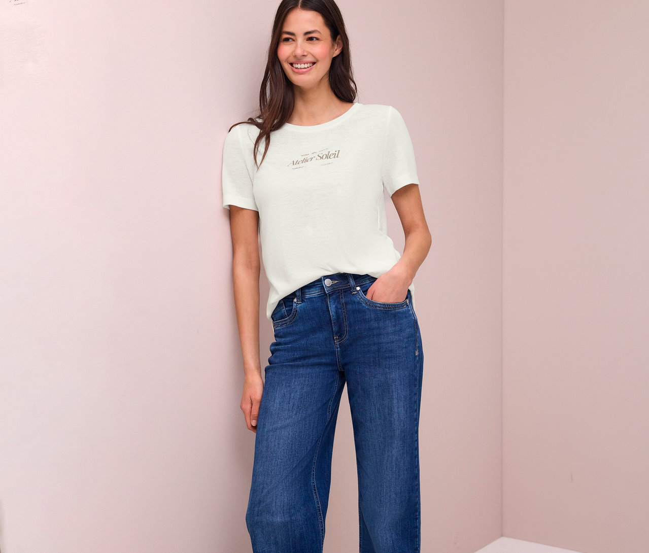 Lächelnde Frau mit Jeans und T-Shirt mit Aufdruck "Atelier Soleil" lehnt an einer Wand.