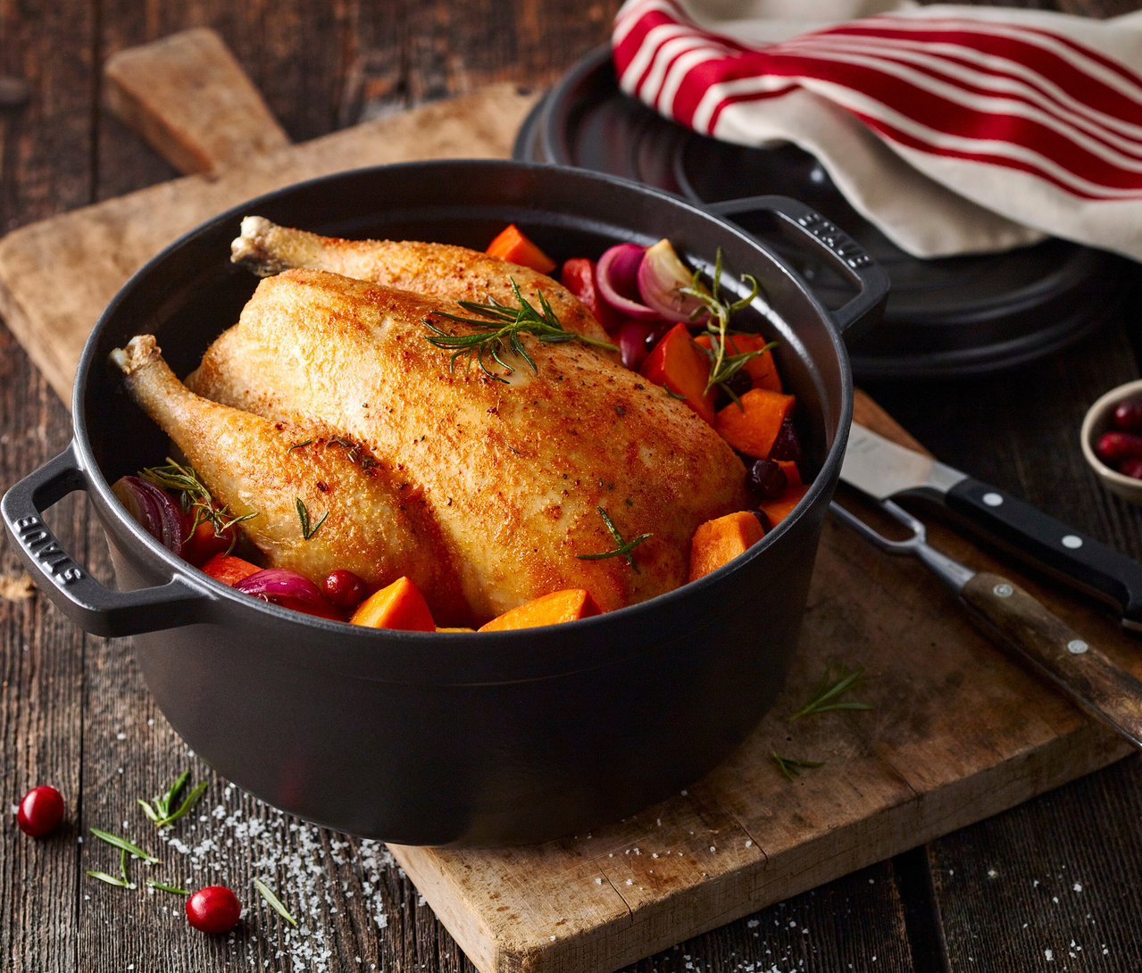 Ganze Brathähnchen in schwarzer STAUB »Cocotte 24« (ca. 3,7 l) mit Gemüse auf Holzbrett.