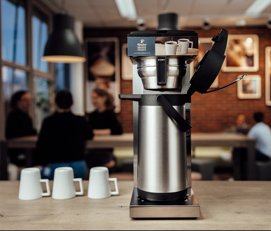 Silberfarbene Tchibo Businessline Profi-Filterkaffeemaschine ohne Kanne auf einem Holztisch mit drei Tassen und Personen im Hintergrund.