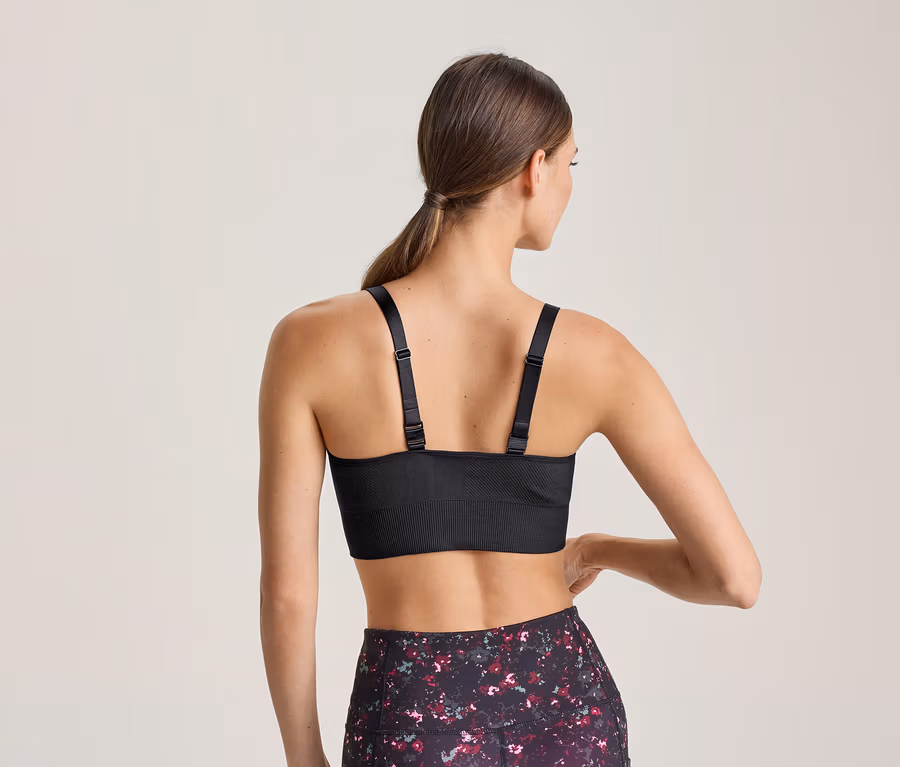 Frau von hinten, die einen schwarzen Sport-BH mit verstellbaren Trägern und gemusterte Leggings trägt. Ihre linke Hand ruht auf ihrer Hüfte.