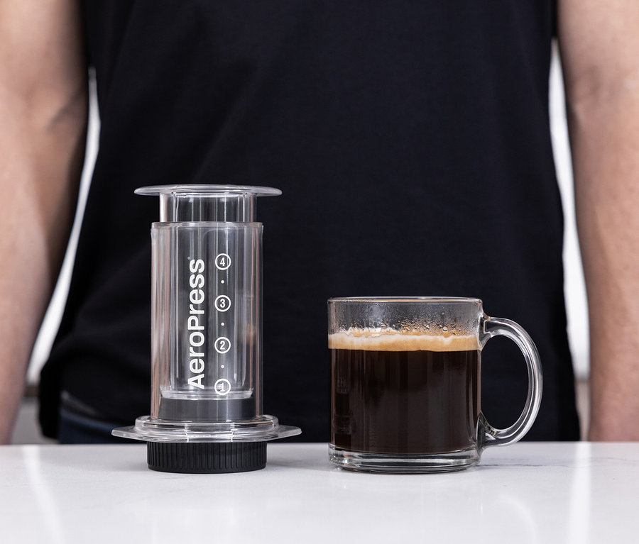 Eine transparente AeroPress Coffee Maker »Clear« steht neben einer Tasse Kaffee.