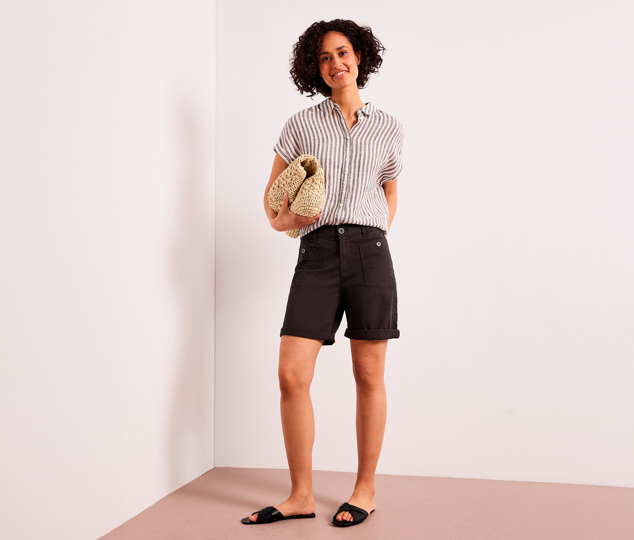Eine lächelnde Frau mit lockigem Haar steht da und hält eine geflochtene Clutch. Sie trägt ein gestreiftes Hemd, braune Street One Bermuda »Yulius« und schwarze Sandalen.