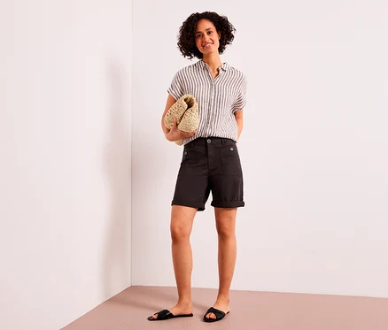 Eine lächelnde Frau mit lockigem Haar steht da und hält eine geflochtene Clutch. Sie trägt ein gestreiftes Hemd, braune Street One Bermuda »Yulius« und schwarze Sandalen.
