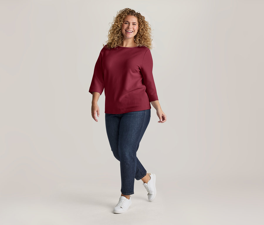Lachende Frau mit lockigem Haar, die ein weinrotes Oberteil, dunkelblaue Jeans und weiße Sneaker trägt und nach vorne geht.