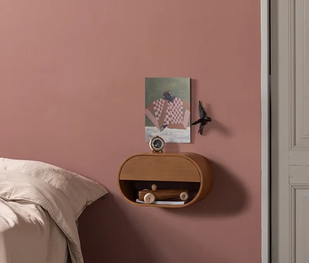 Schlafzimmer mit altrosafarbener Wand, Bett mit heller Bettwäsche und hellem Fischgrätparkett. Ein schwebender Nachttisch aus Holz trägt einen Wecker und ein Holzspielzeugauto. Ein abstraktes Gemälde und ein Dekovogel sind an der Wand.