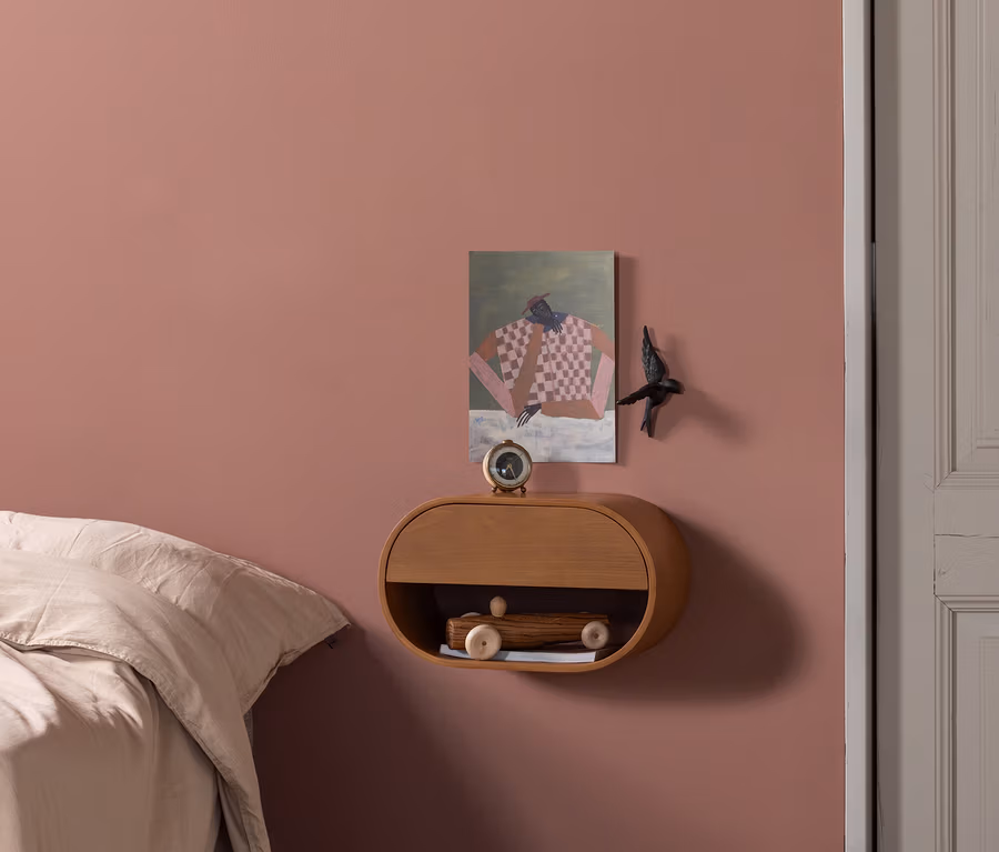 Schlafzimmer mit altrosafarbener Wand, Bett mit heller Bettwäsche und hellem Fischgrätparkett. Ein schwebender Nachttisch aus Holz trägt einen Wecker und ein Holzspielzeugauto. Ein abstraktes Gemälde und ein Dekovogel sind an der Wand.