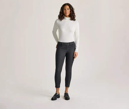 Eine Frau mit dunklem, lockigem Haar steht da und trägt ein weißes, langärmeliges Rollkragenshirt und dunkelgraue Treggings - Fit »Nelly«, mit den Händen in den Taschen.