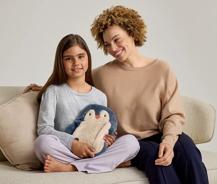 Eine erwachsene Frau und ein junges Mädchen sitzen lächelnd auf einem Sofa. Das Mädchen links hält ein blau-beiges Pinguin-Stofftier. Die Frau rechts hat ihren Arm um das Mädchen gelegt.