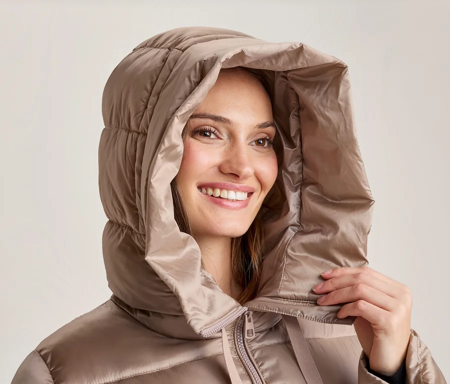 Lächelnde Frau in einer beigen Steppjacke mit hochgezogener Kapuze, die eine Hand am Kragen hält.