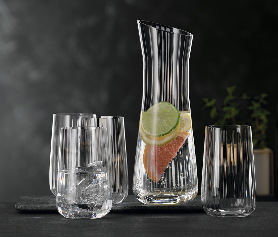 Eine klare Glaskaraffe mit Wasser, Limetten- und Grapefruitscheiben, umgeben von mehreren klaren Spiegelau Longdrinkgläsern #Lifestyle#, eines davon mit Eis und Wasser gefüllt, auf dunklem Untergrund.