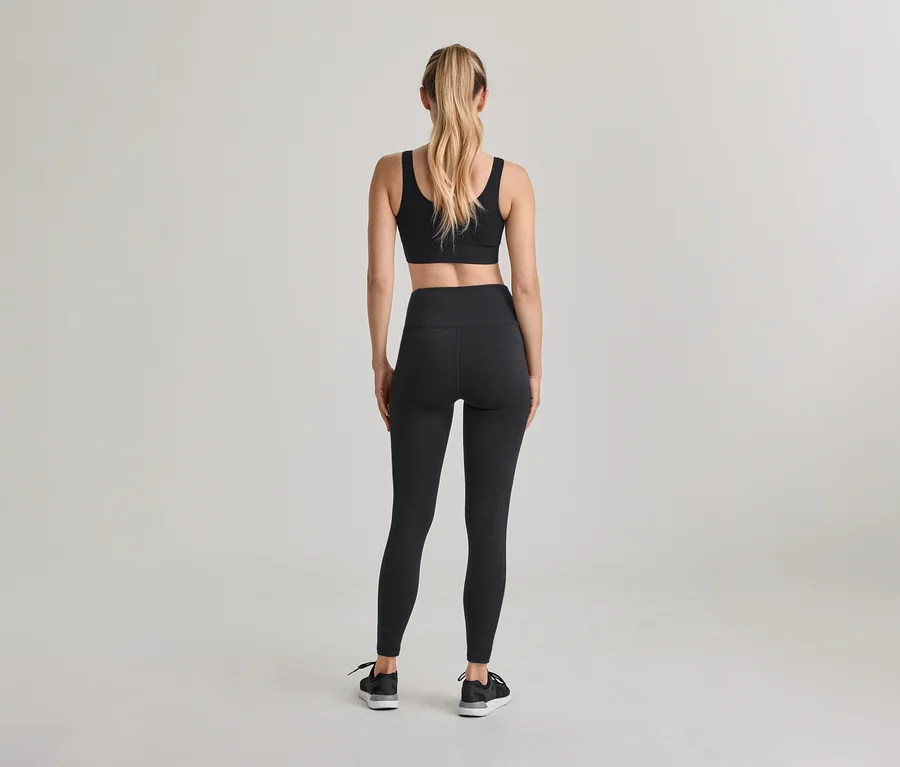 Frau von hinten, trägt schwarzen Sport-BH, schwarze High-Waist-Leggings und schwarze Sneaker. Ihr blondes Haar ist zu einem Pferdeschwanz gebunden.