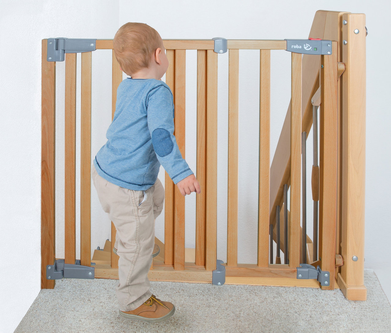 Ein Kleinkind in blauem Shirt und beiger Hose steht vor einem hölzernen roba Treppenschutzgitter »Safety Up« und blickt zur Absperrung am oberen Ende einer Treppe.