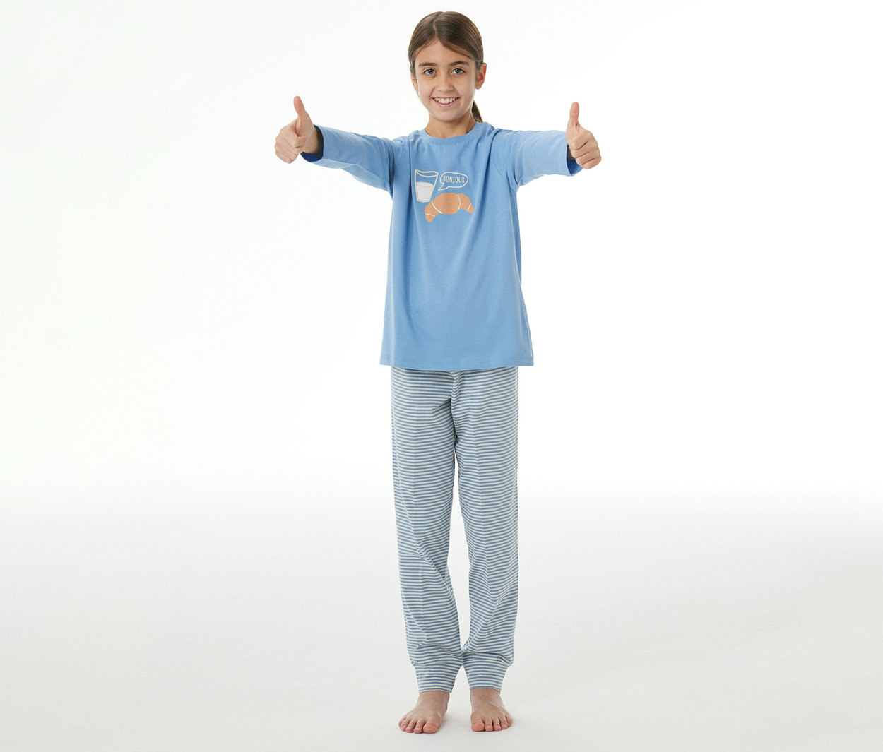 Lächelndes junges Mädchen im SCHIESSER Kinder-Pyjama, blaues Oberteil mit Milchglas- und Croissant-Motiv und gestreifter Hose, zeigt beide Daumen hoch.