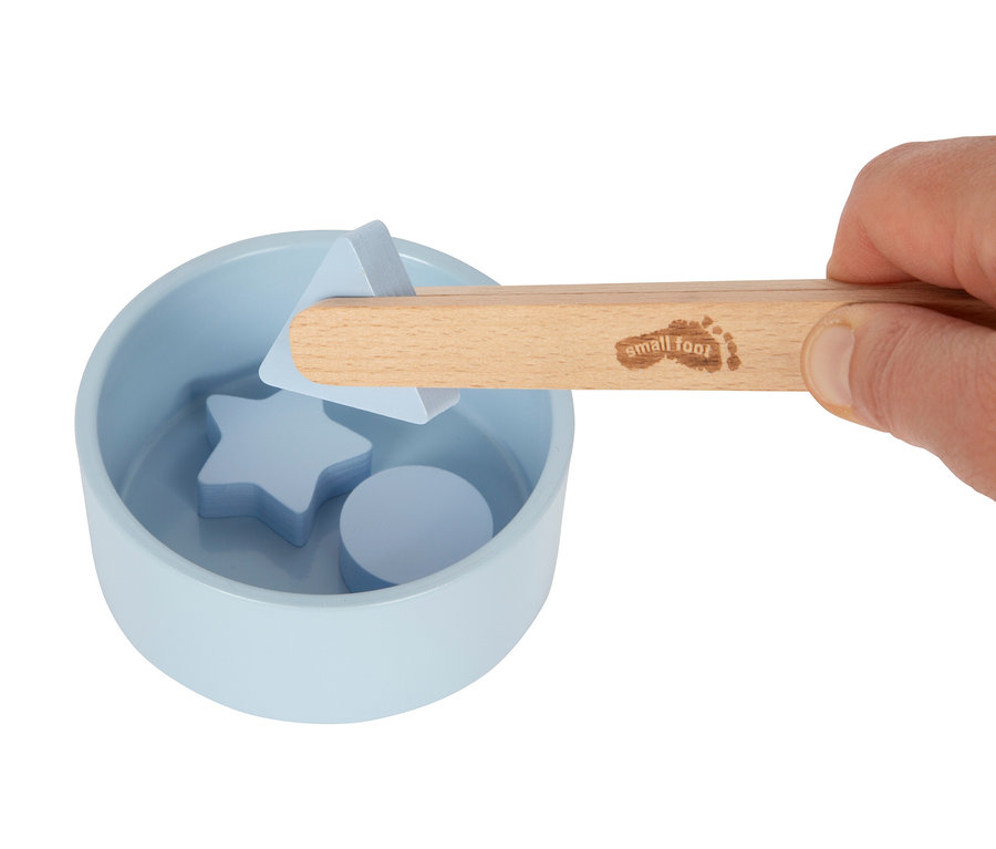 Eine Hand nimmt mit einer Holzzange ein hellblaues Holzdreieck aus einer hellblauen Schale. Eine hellblaue Holzstern- und Kreisform sind ebenfalls in der Schale. Die Zange trägt das "small foot" Logo.
