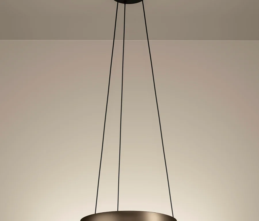 Eine moderne, kreisförmige Pendelleuchte mit dunklem Metallfinish hängt an drei schwarzen Kabeln von einer hellen Decke. Ein LED-Streifen am Innenrand des Rings strahlt warmes Licht nach unten ab.