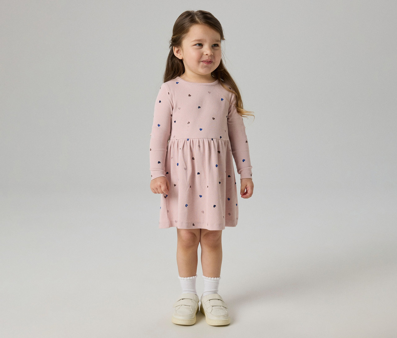 Kleines Mädchen mit langen braunen Haaren, das ein rosa Langarmkleid mit Herzmustern, weiße Rüschensocken und helle Schuhe trägt. Sie steht lächelnd und blickt nach rechts.